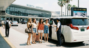 成田空港のターミナルビル前で、夏服を着た4人の若い女性グループが、白いトヨタ・アルファードの鍵を制服を着たバレーパーキングの係員に手渡している様子。背景には「NARITA AIRPORT」の文字と「バレーパーキング受付」のデジタル看板が見える晴れた日の光景。